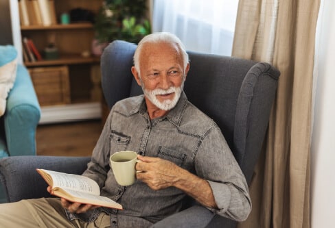 man sitting in a high backed chair reading a book while enjoying some coffee